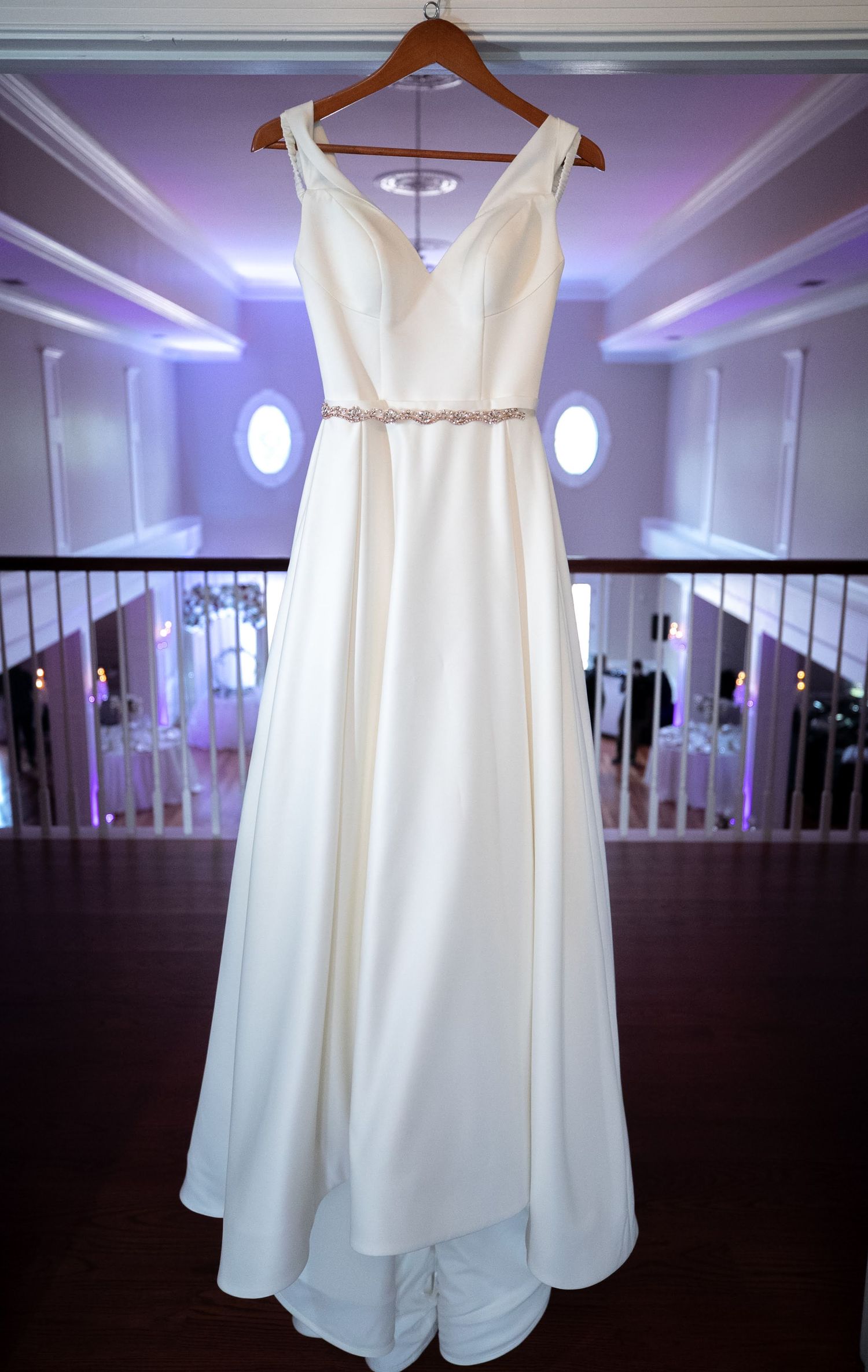 Elegant white A-line wedding gown with jeweled belt hanging on a wooden hanger indoors.