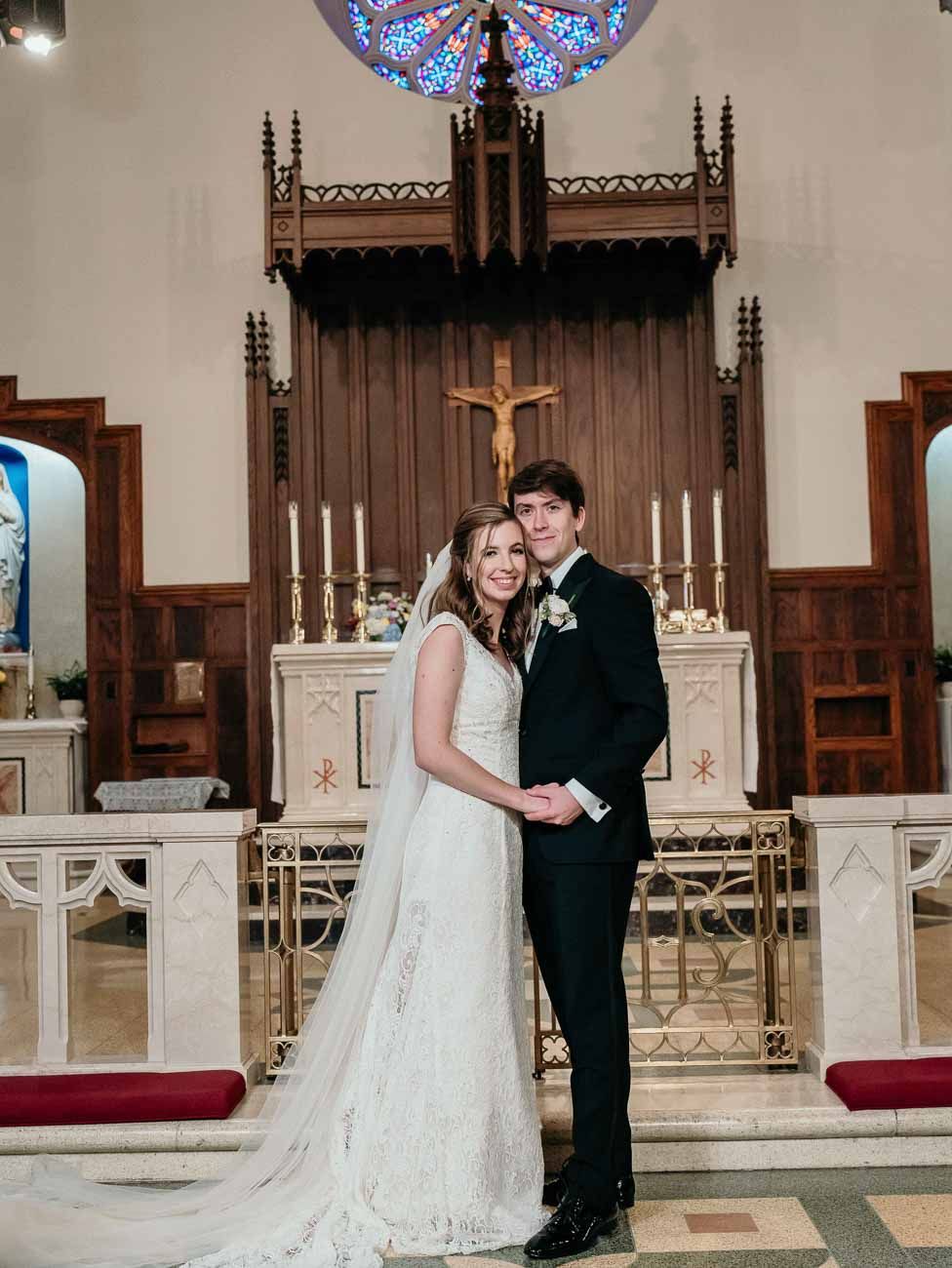Newlyweds pose together at the altar of a Catholic church with candles and a crucifix backdrop.