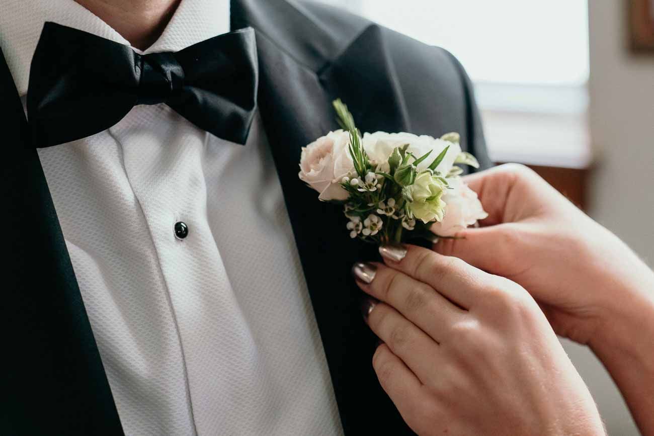 Hands pinning white rose boutonniere on groom in black tuxedo with bow tie.