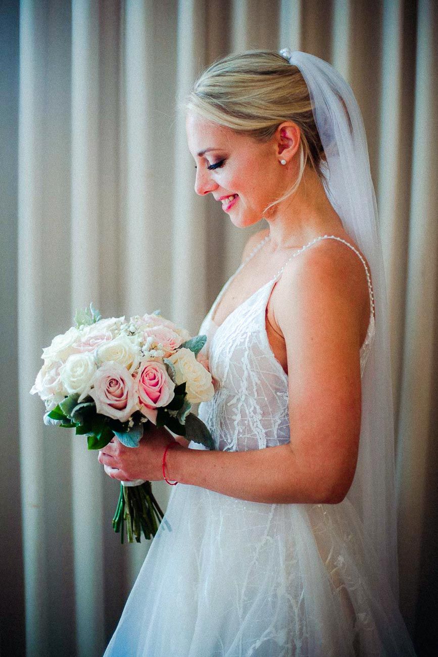 Elegant blonde bride in white lace gown holding pink and white rose bouquet with veil.