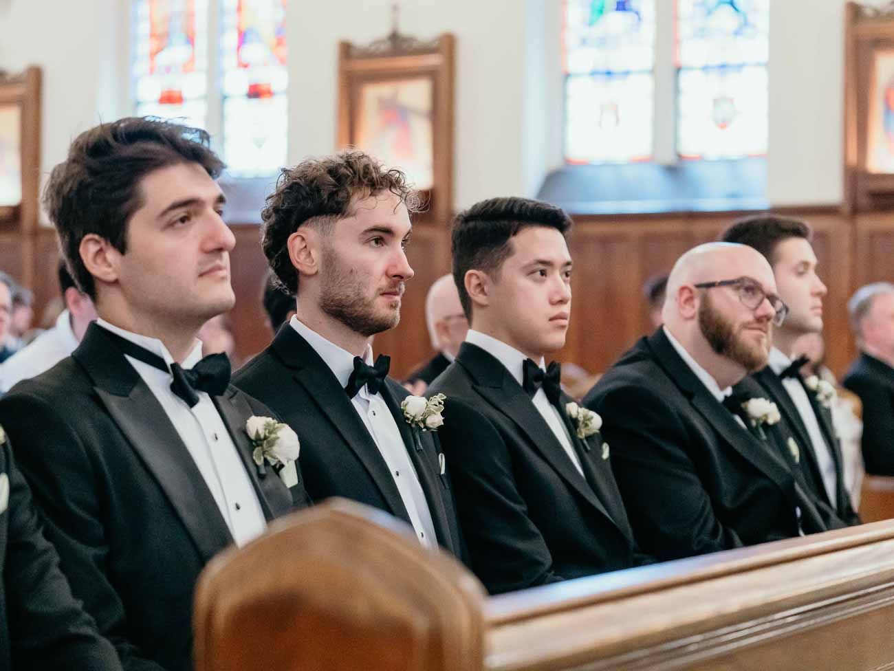 Four groomsmen in black tuxedos with boutonnieres seated in church pews during a wedding.