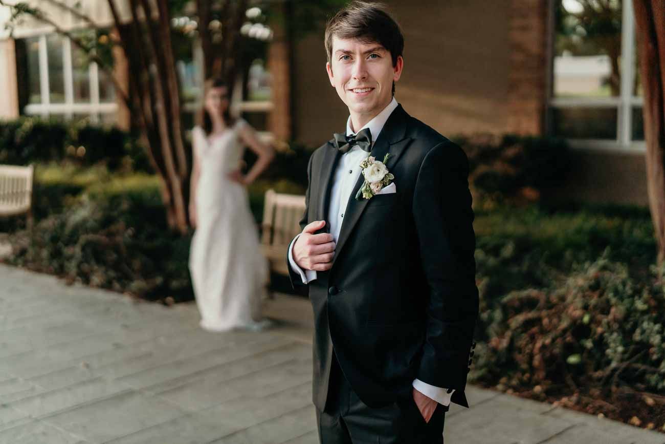 Groom in black tuxedo smiling confidently outdoors with bride in white lace gown visible in background.