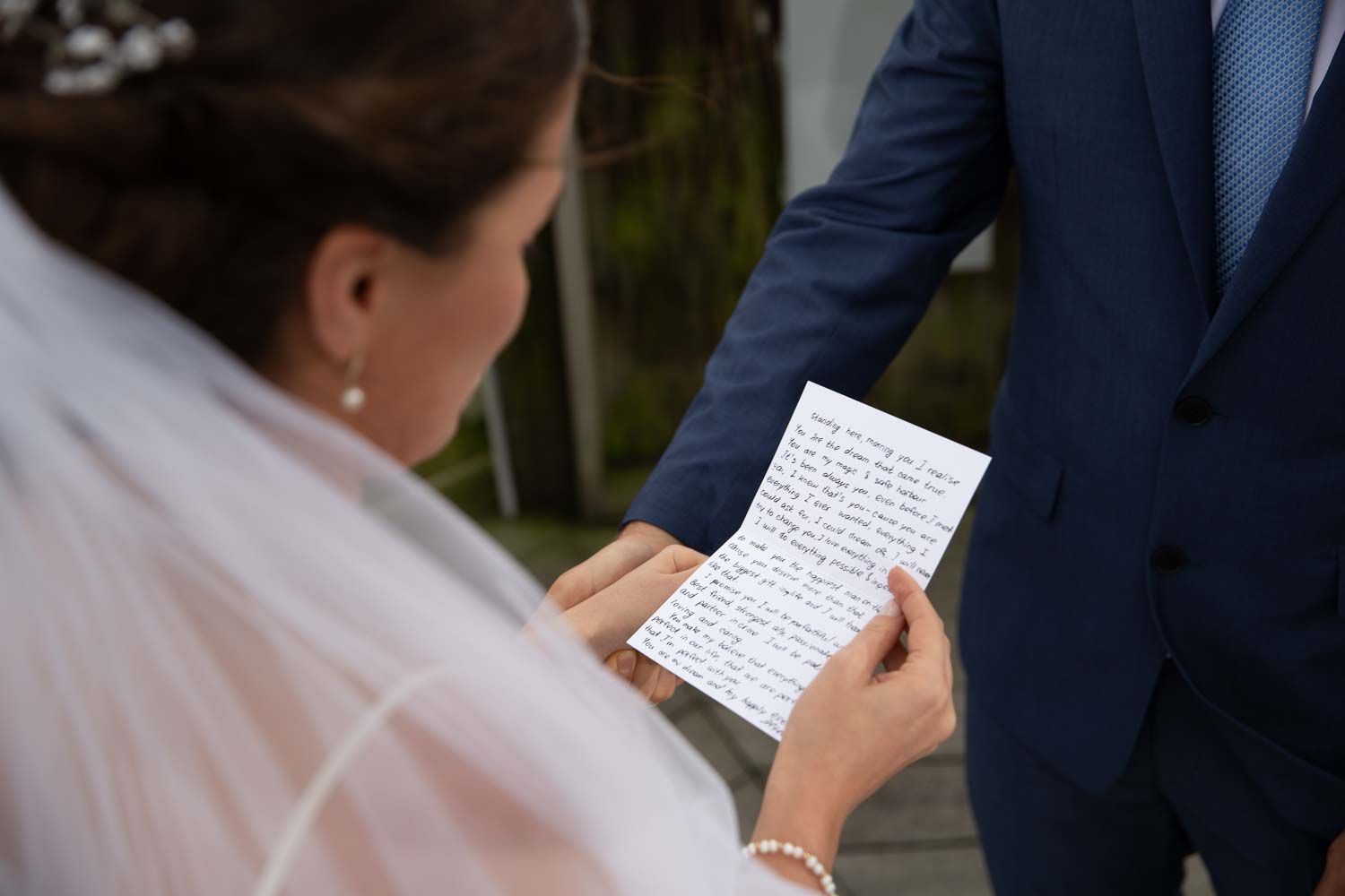 Bride reads handwritten wedding vows letter while groom in blue suit stands beside her outdoors.