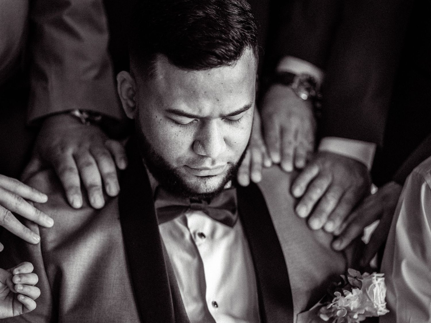 Black and white photo of a groom in bow tie with eyes closed, hands placed on his shoulders in prayer.