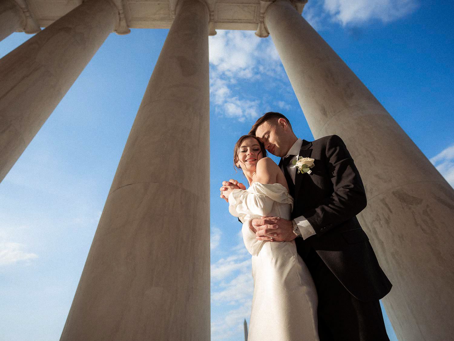 Bride and groom pose intimately beneath grand marble columns with a bright blue sky backdrop.