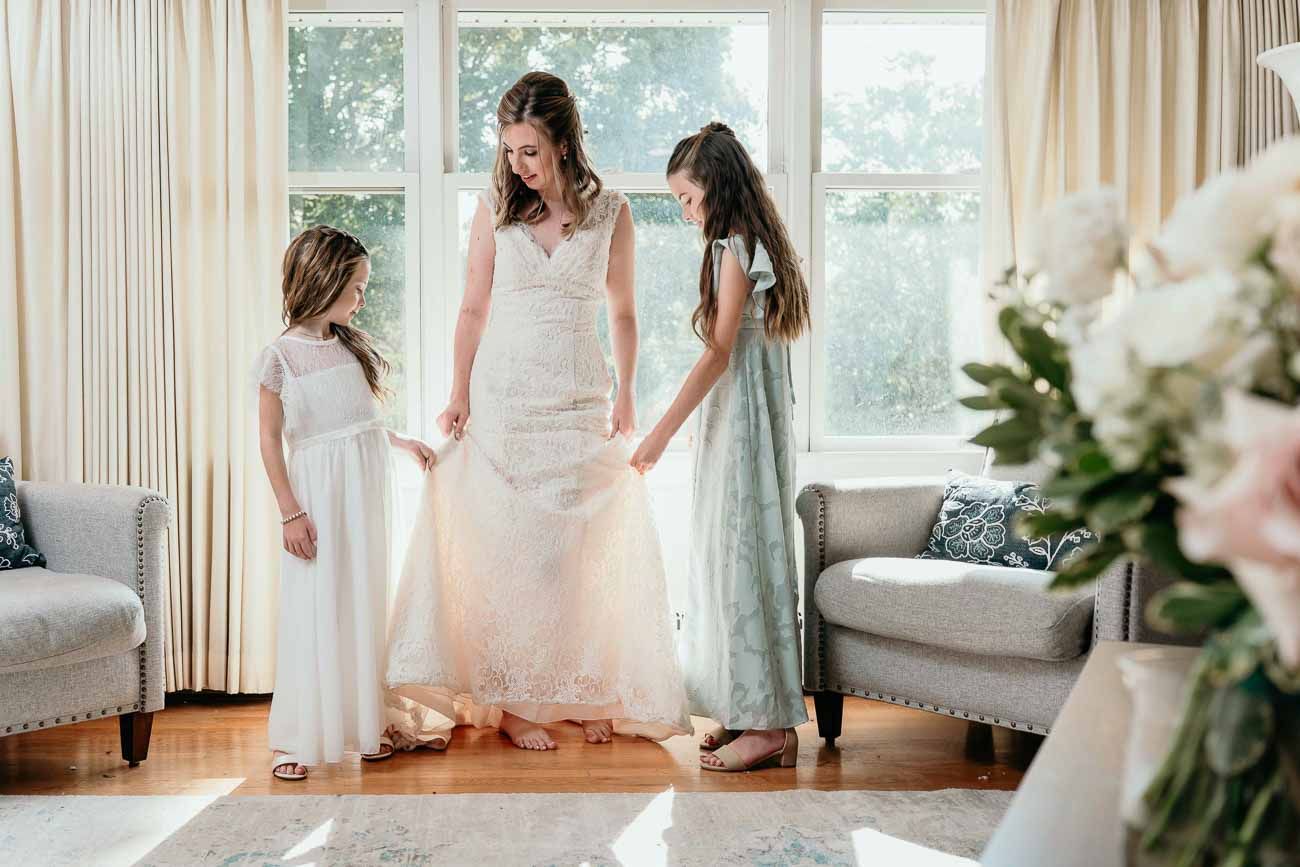 Bride in white lace wedding gown holds hands with two young flower girls in elegant dresses indoors.