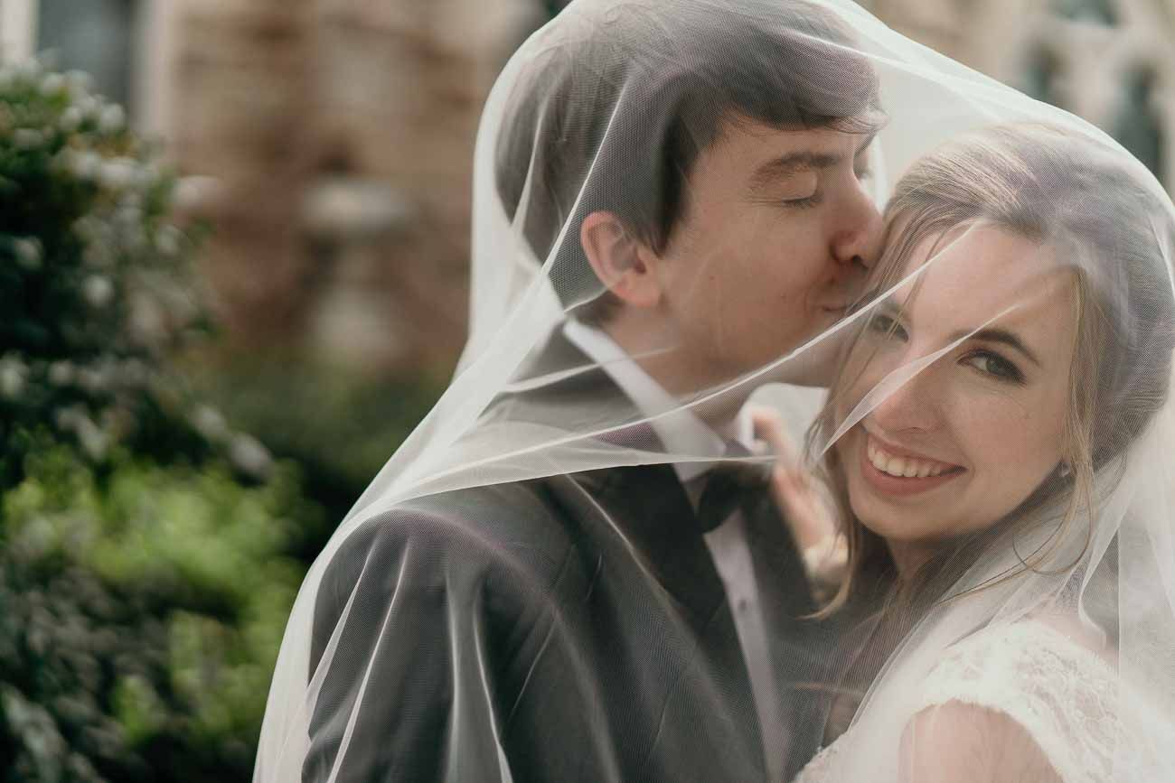 Groom tenderly kisses bride's cheek as she smiles, both draped under her flowing white veil.