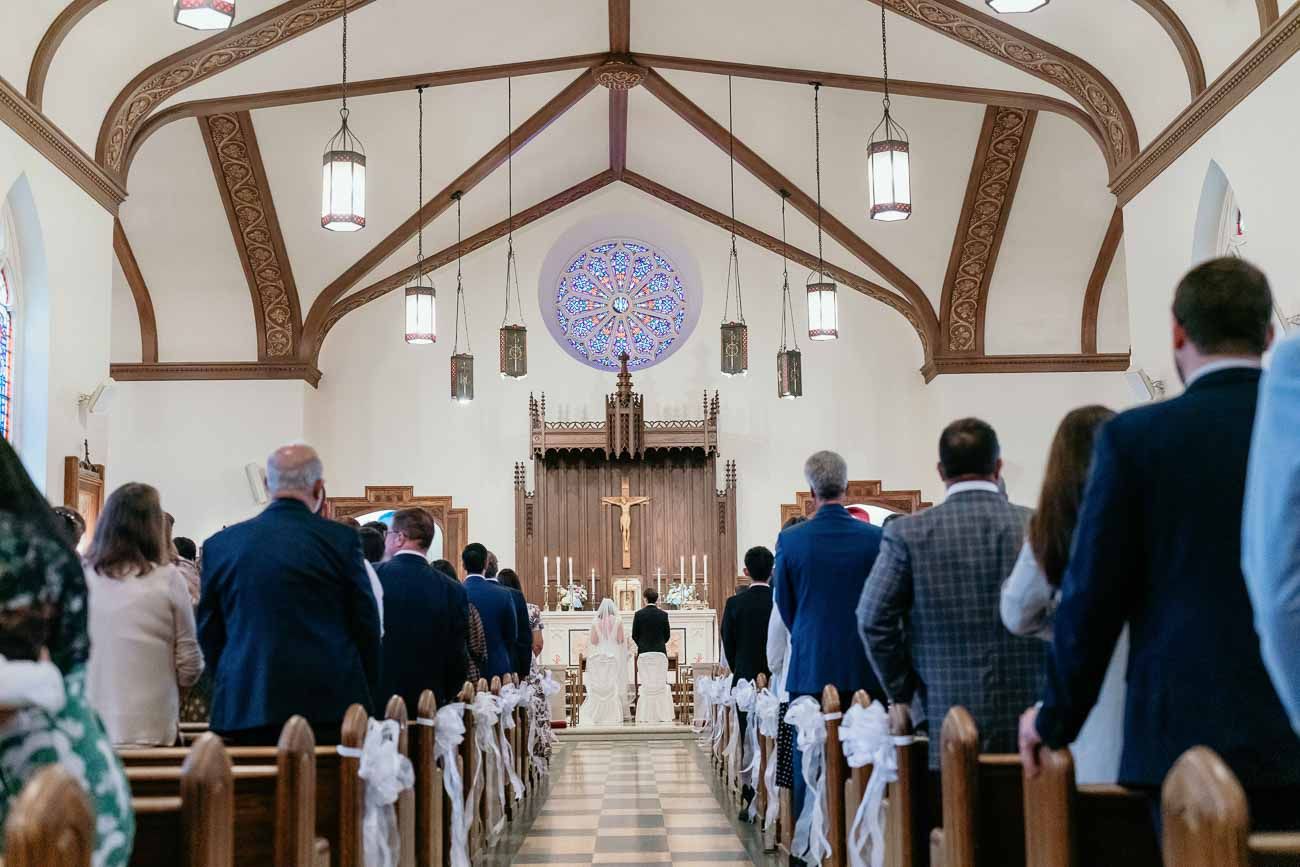Wedding guests stand in church pews during a Catholic wedding ceremony with a rose window altar backdrop.