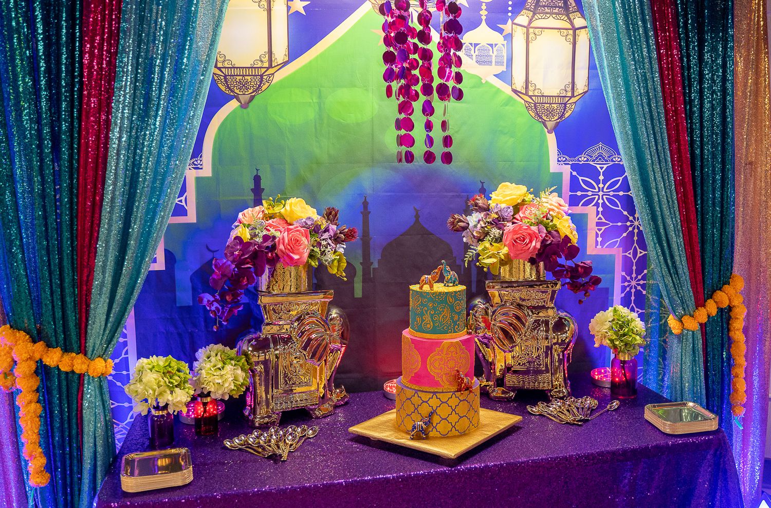 Moroccan-themed dessert table with ornate birdcages, colorful cake, and purple sequin backdrop with lantern motifs.