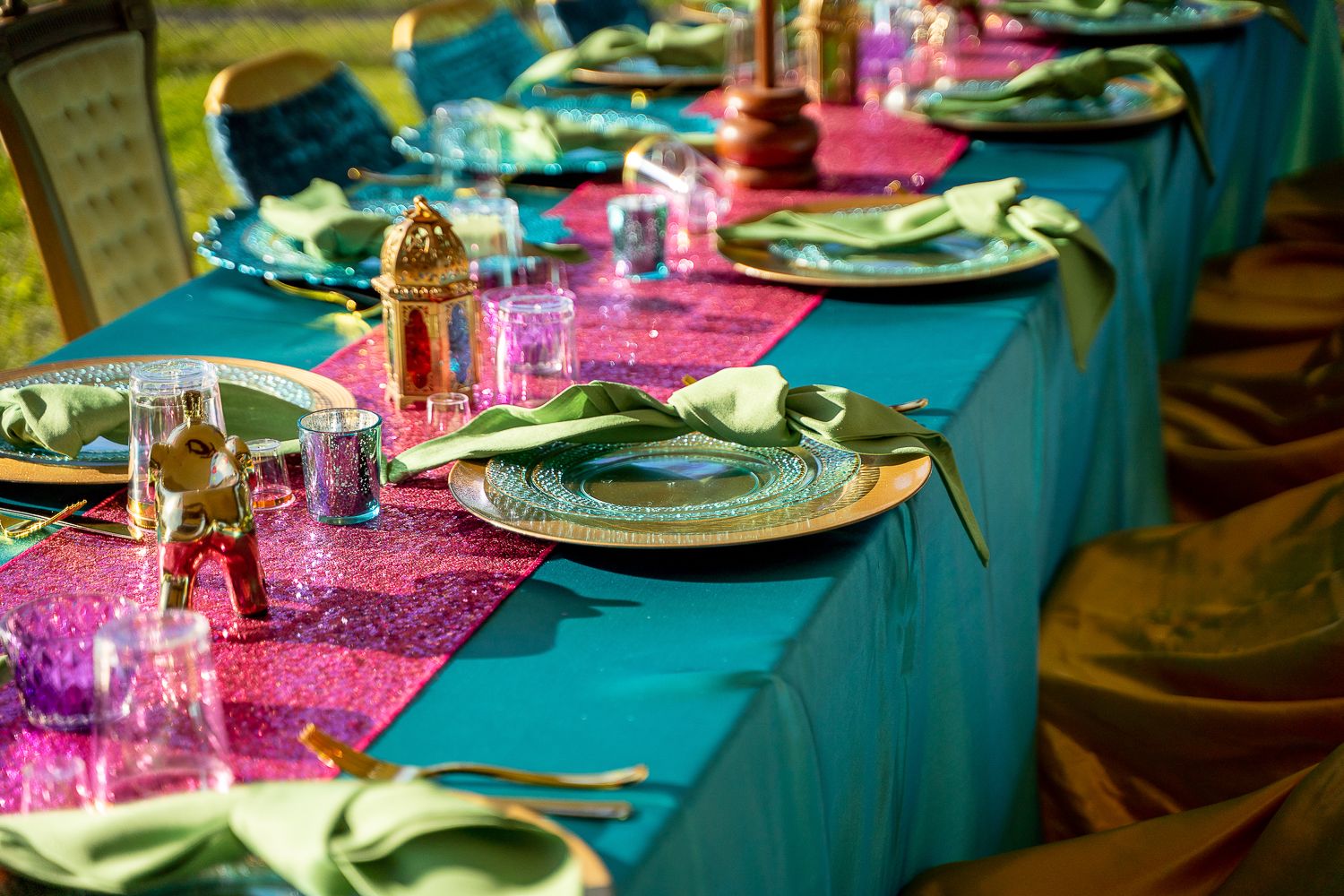 Elegant Arabian Nights table setting with teal tablecloth, pink sequin runner, gold chargers, and green napkins.