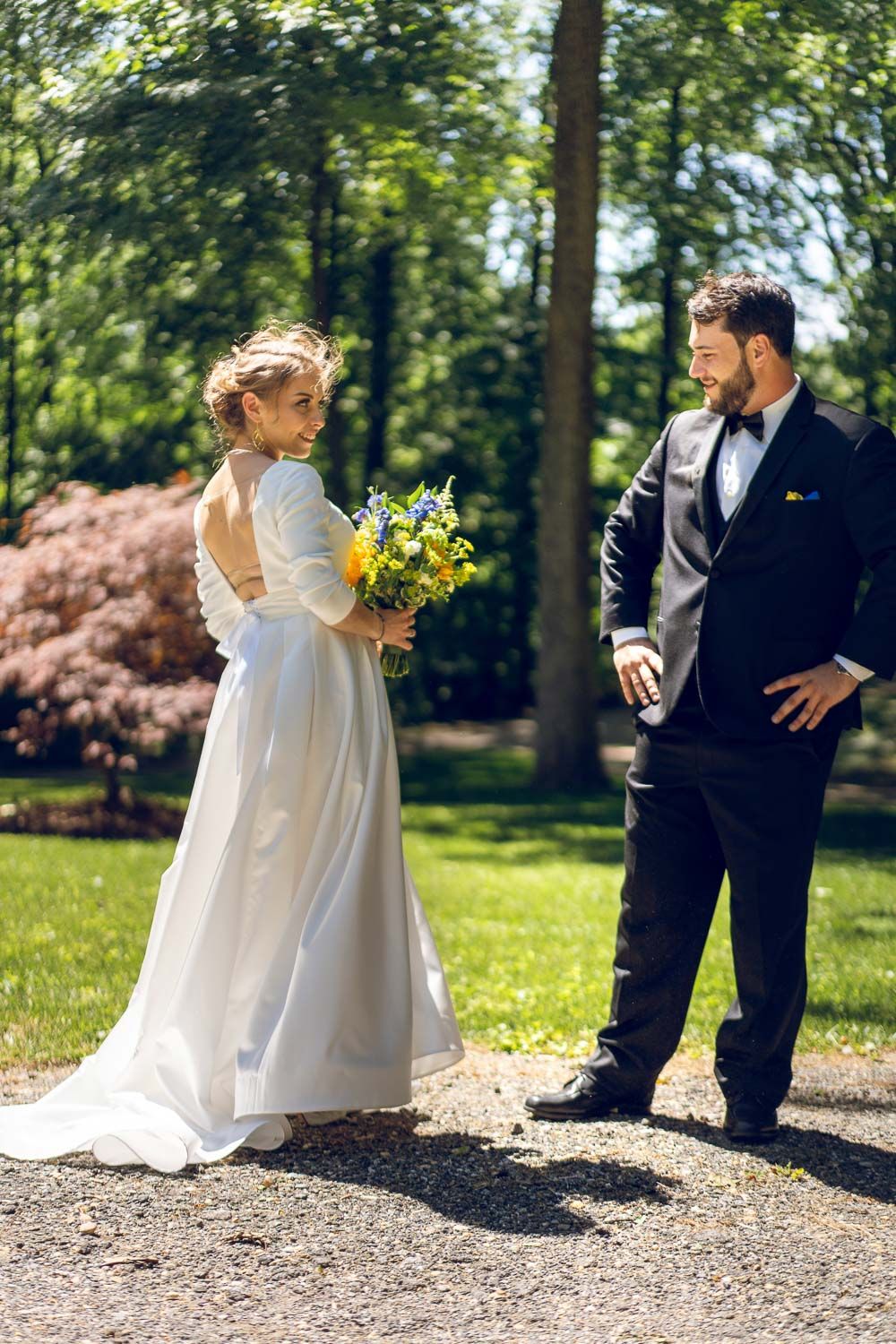 Bride in backless white gown glancing back at smiling groom on garden path outdoors.