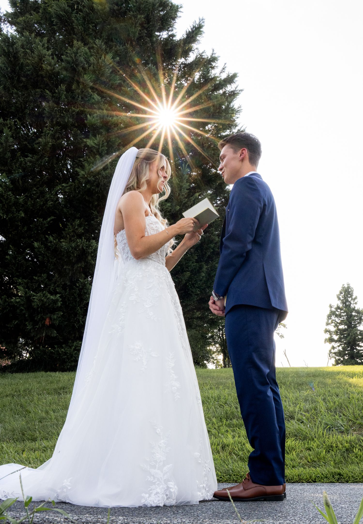 Bride reads vows to groom outdoors, sun burst through evergreen trees, white lace gown and navy suit.