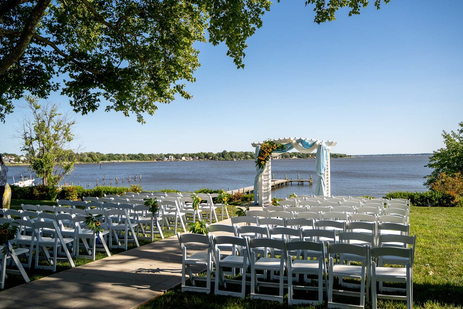 Elegant outdoor wedding ceremony setup with white chairs facing a floral arch by the waterfront.