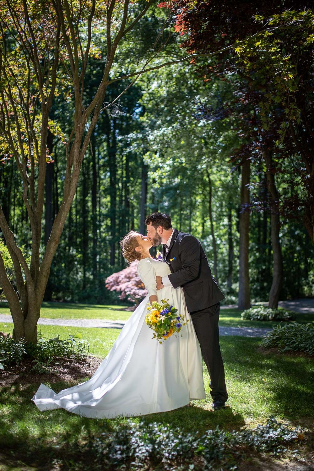 Bride and groom sharing romantic kiss under lush green trees with colorful wildflower bouquet.