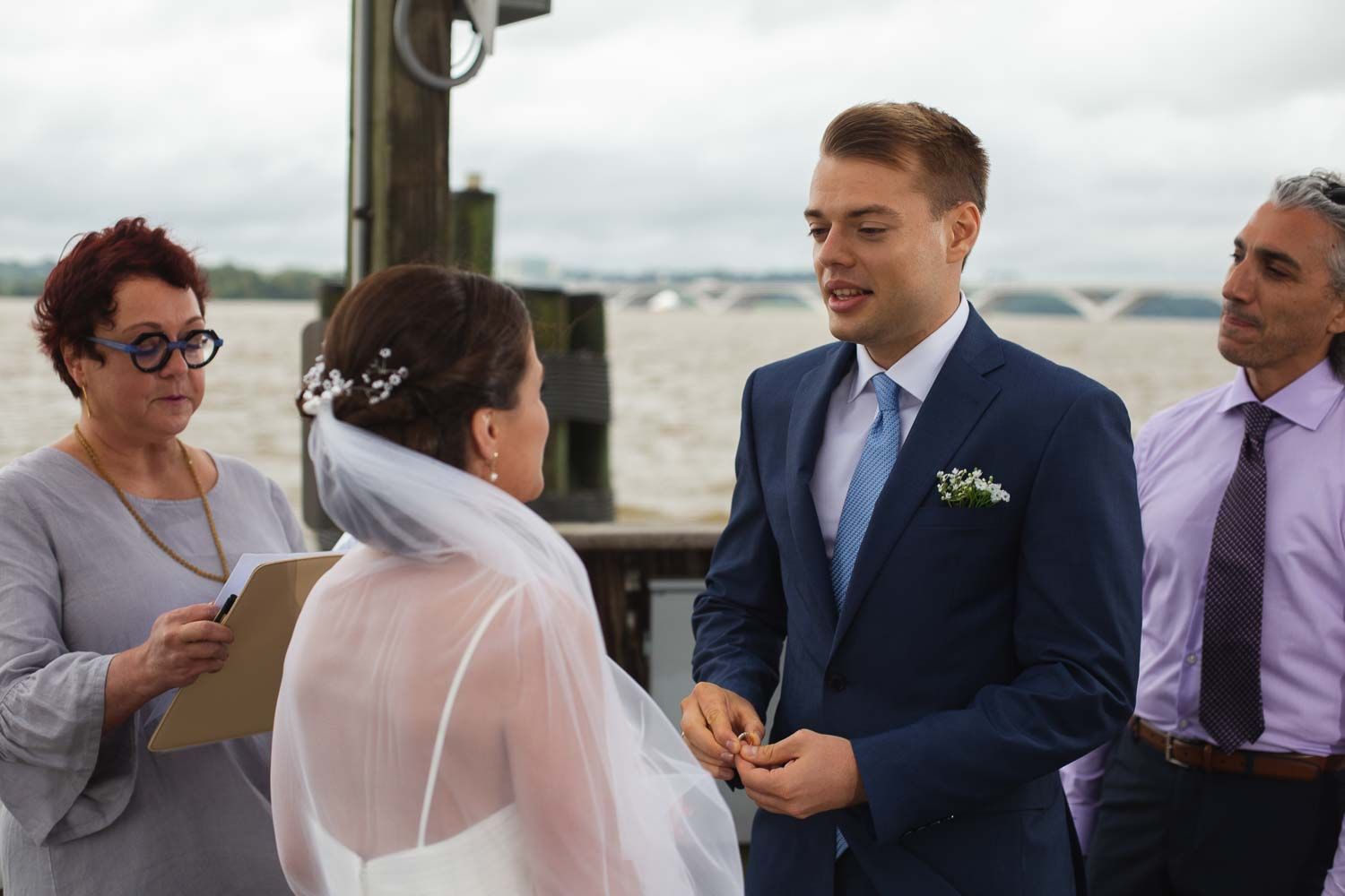 Smiling groom in blue suit holds brides hands during outdoor waterfront wedding ceremony with guests present.