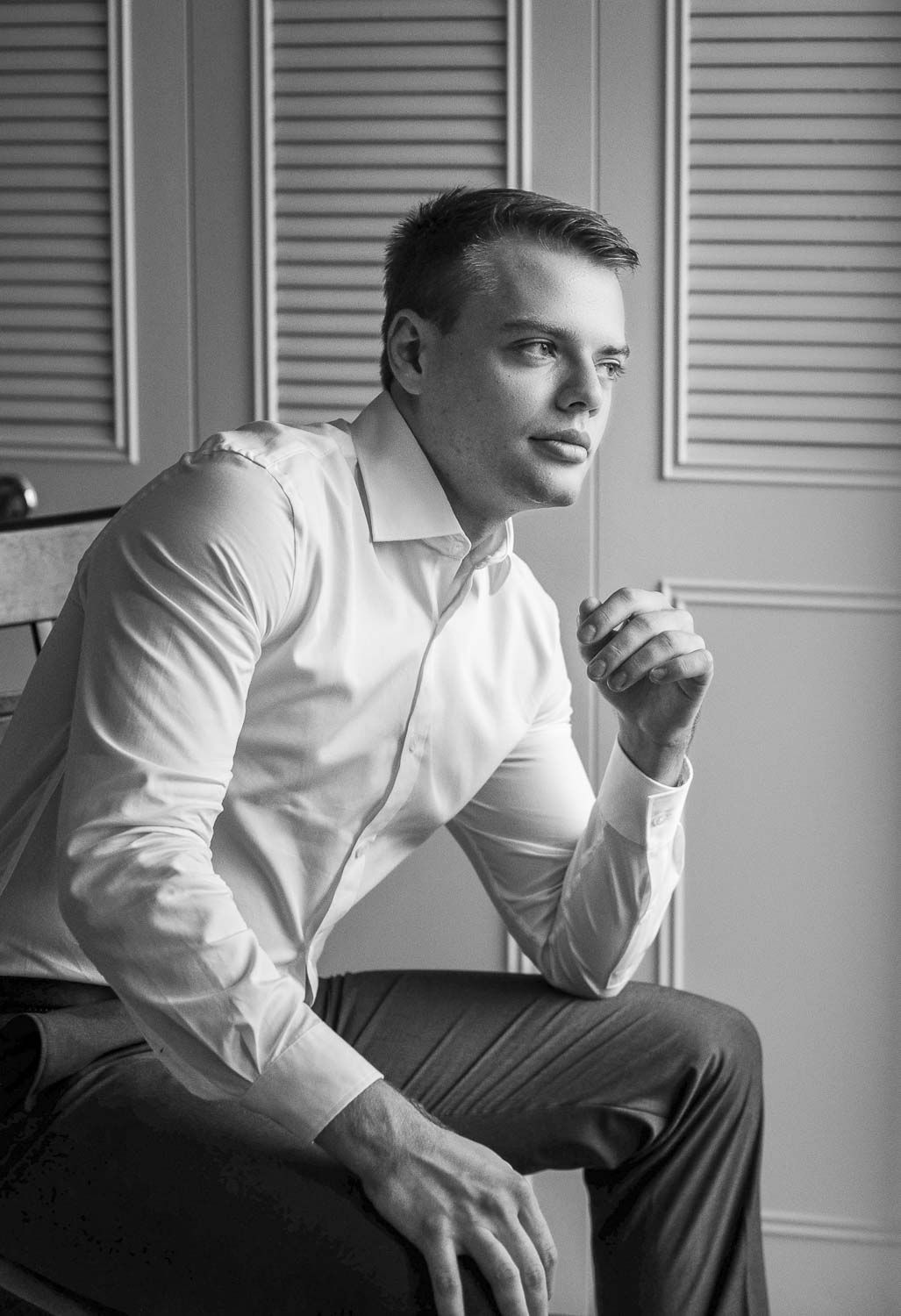 Black and white portrait of a young man in a white dress shirt sitting thoughtfully in an elegant room.