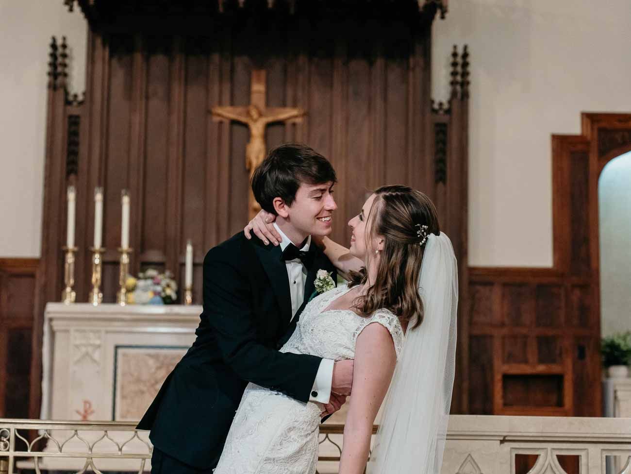 Bride and groom share a romantic moment at the altar of a traditional Catholic church with a wooden crucifix.
