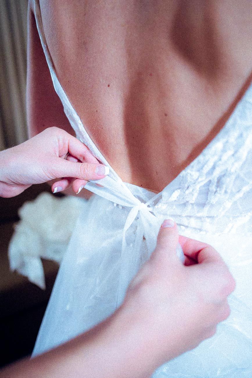 Bridesmaid tying delicate ribbon bow on back of bride's white lace wedding dress.