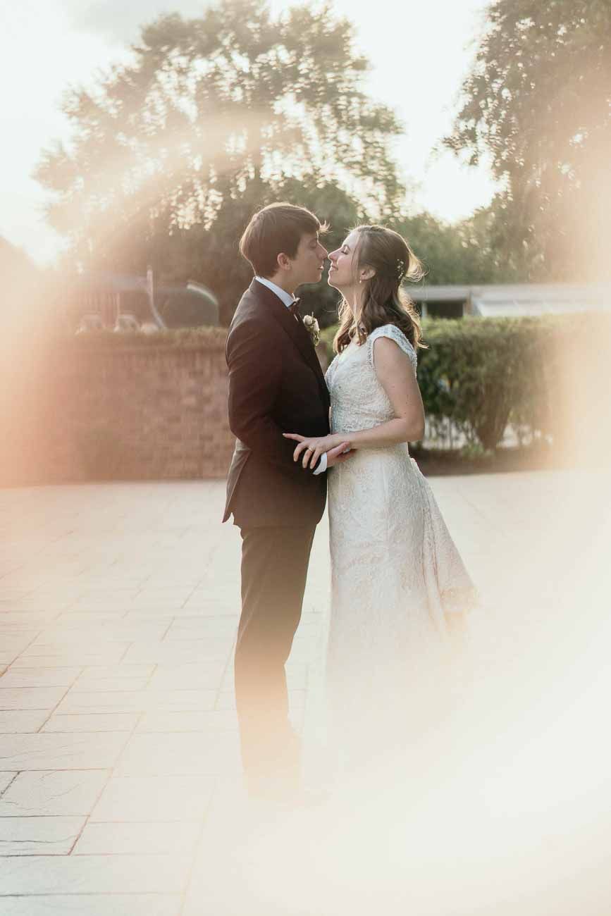 Bride and groom facing each other in a golden sunlit outdoor setting, about to share a kiss.