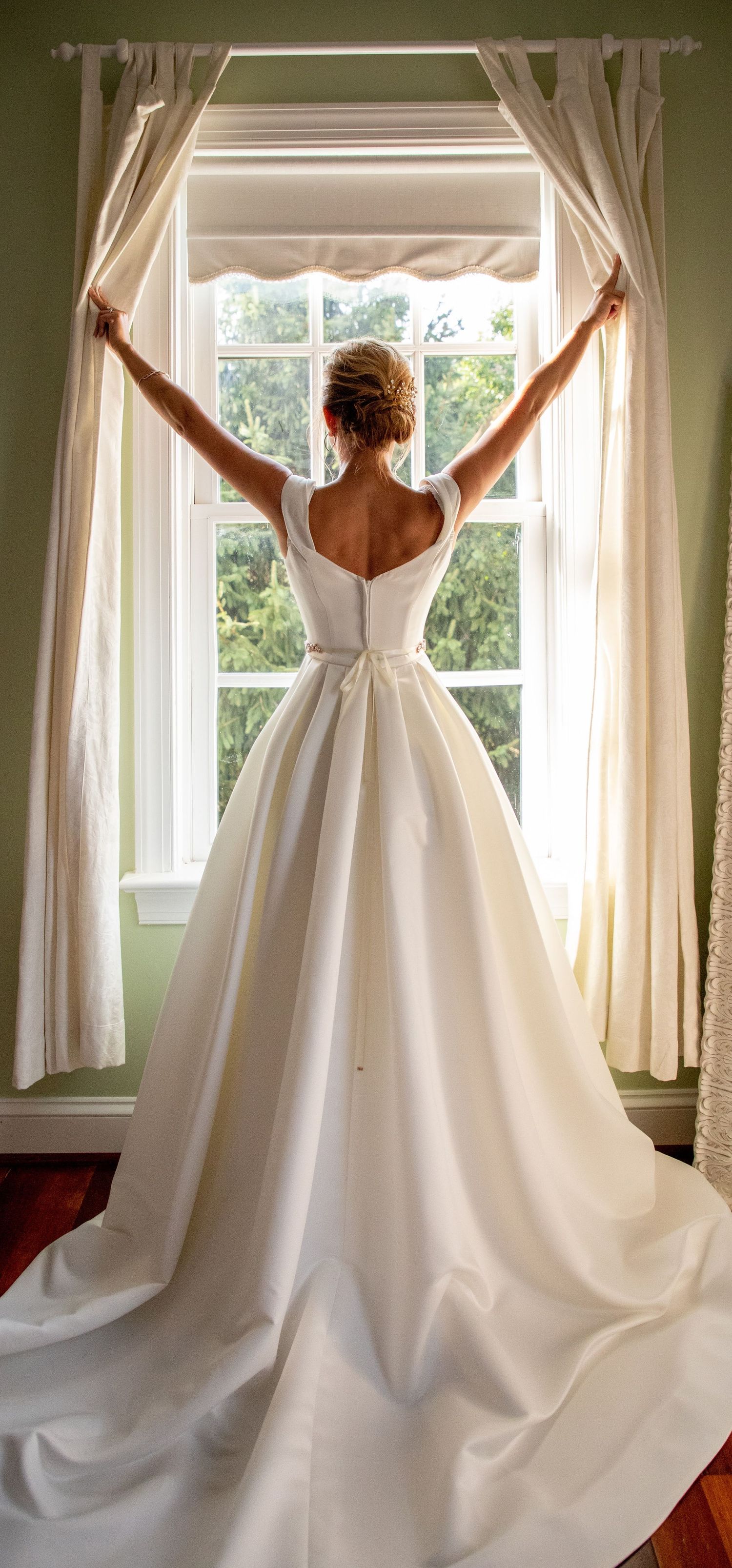 Bride in classic white ball gown with train stands at window, arms outstretched holding curtains.