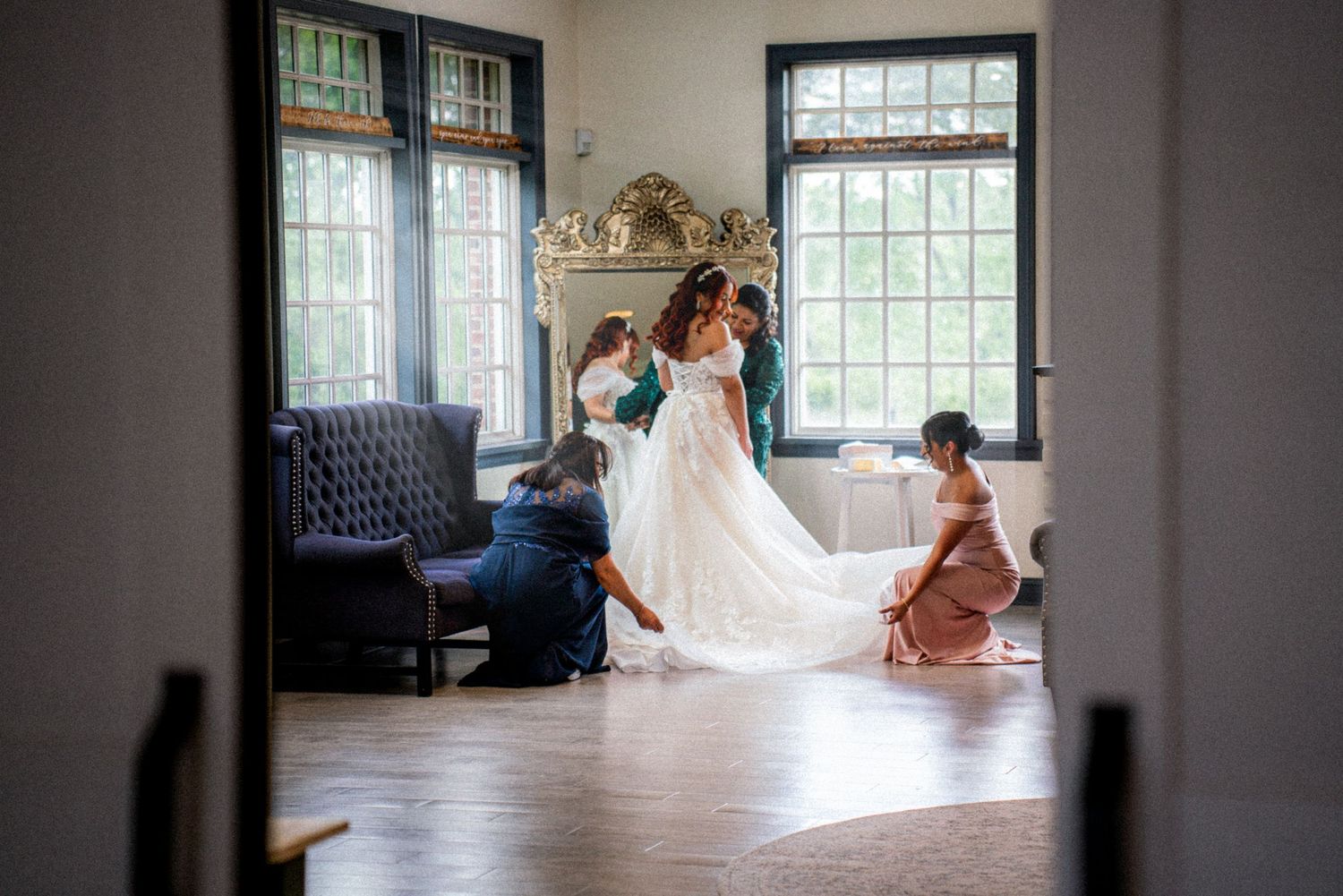 Bride in white gown being helped by bridesmaids in elegant room with large windows and ornate decor.