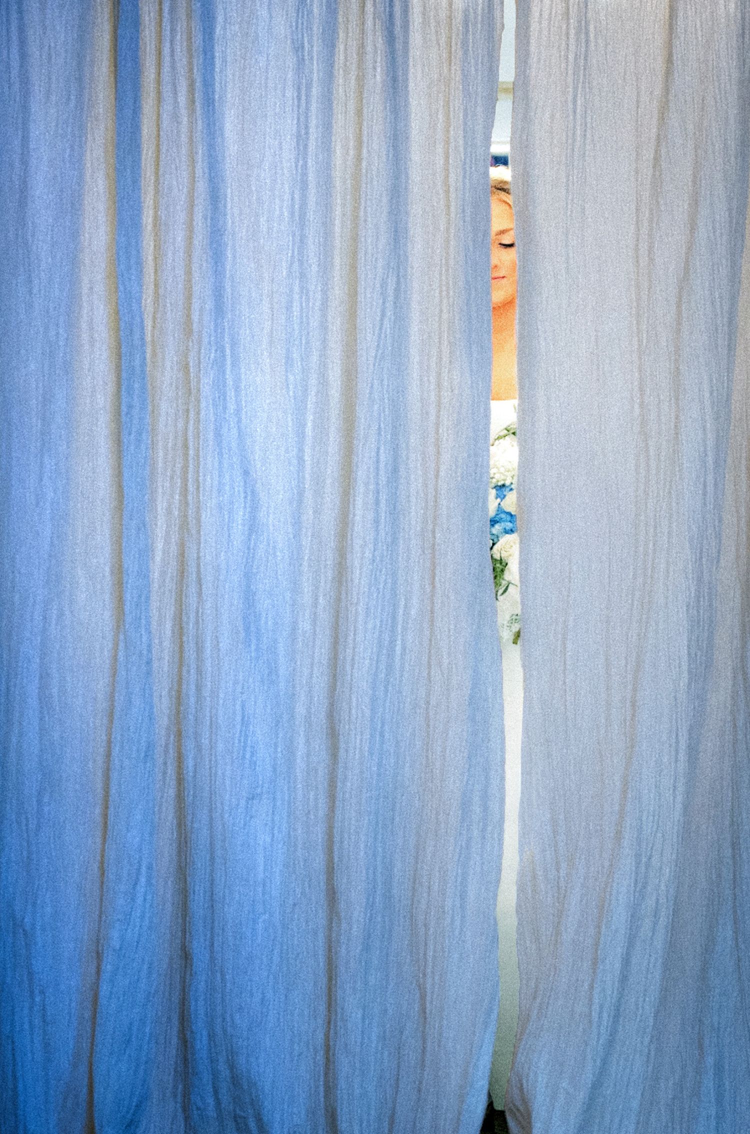 Sheer blue curtains partially open revealing a glimpse of colorful fabric and a sleeping person behind them.