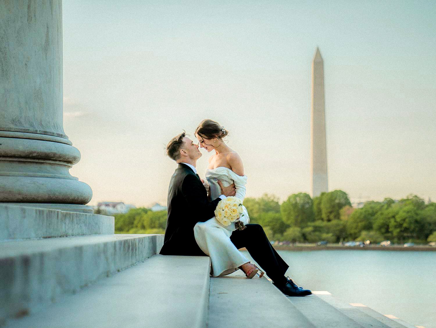 Newlyweds share a tender kiss on stone steps with Washington Monument at dusk.