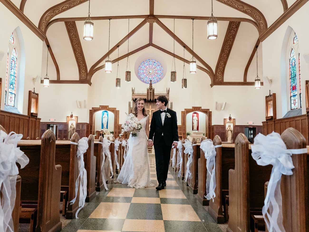 Bride and groom walk down the aisle of a decorated church with white ribbon pews and stained glass windows.