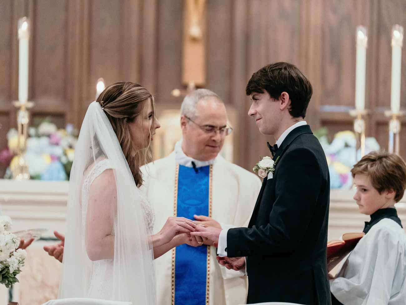 Smiling bride and groom hold hands during ring exchange with priest in Catholic wedding.