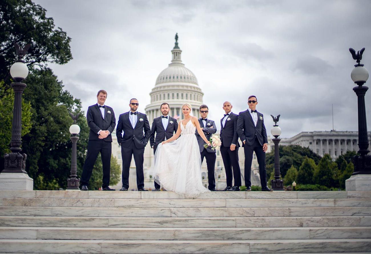Bride in white gown flanked by groomsmen in black tuxedos on Capitol Building steps.