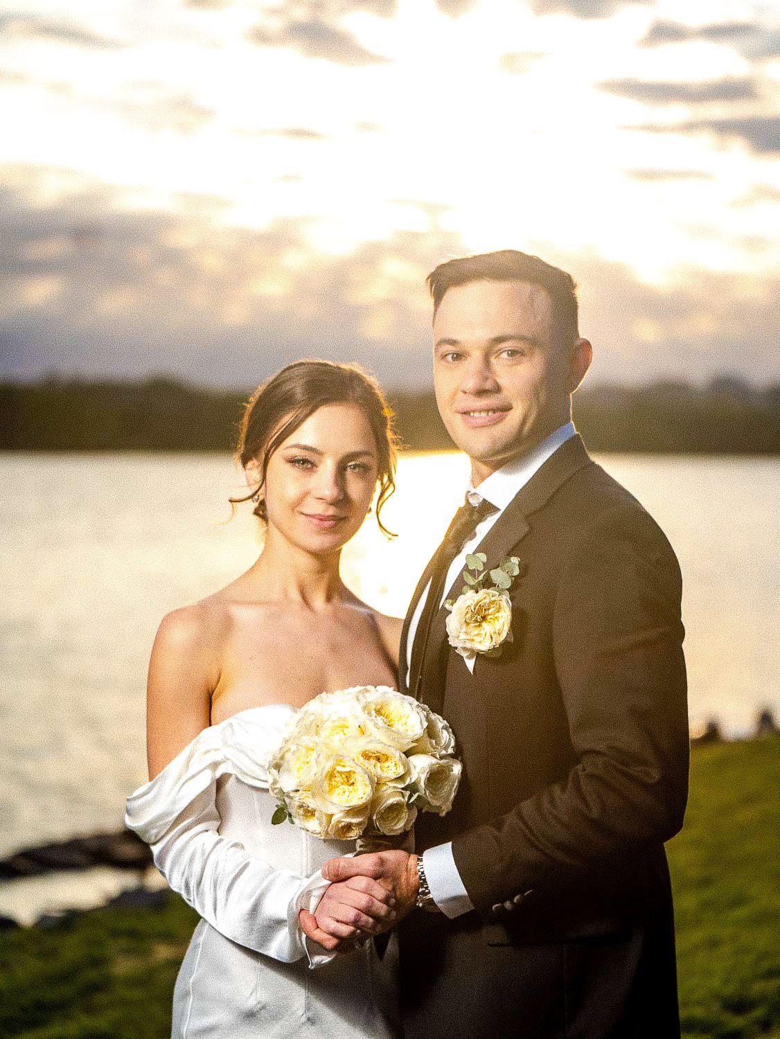 Smiling bride and groom pose with white bouquet in front of a dramatic sunset over the water.