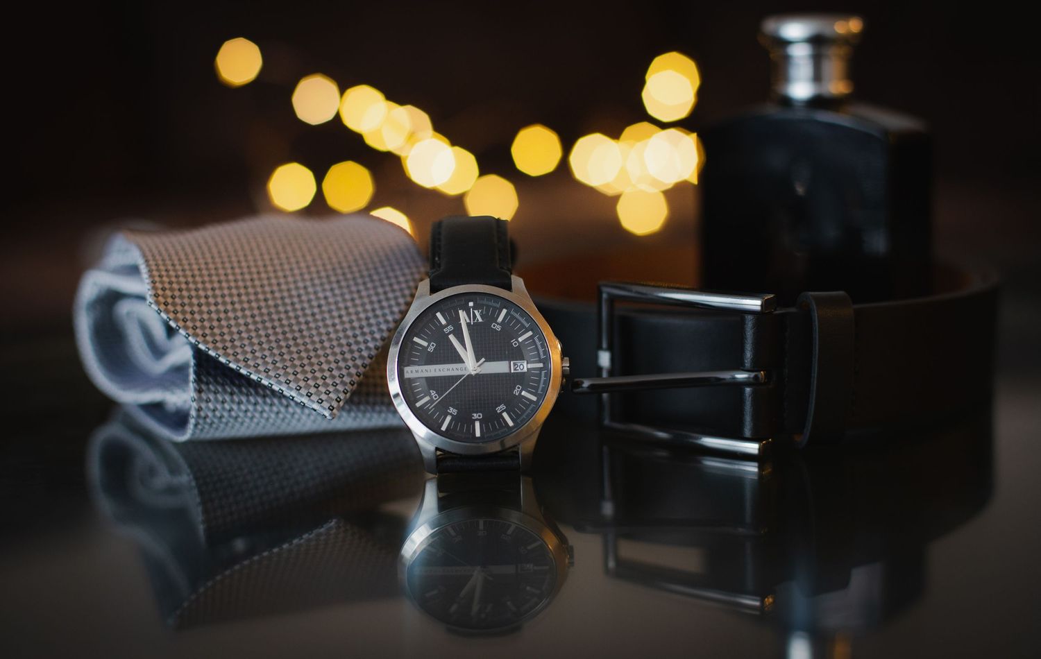 A classic black watch, leather belt, tie, and cologne arranged as groom wedding accessories.
