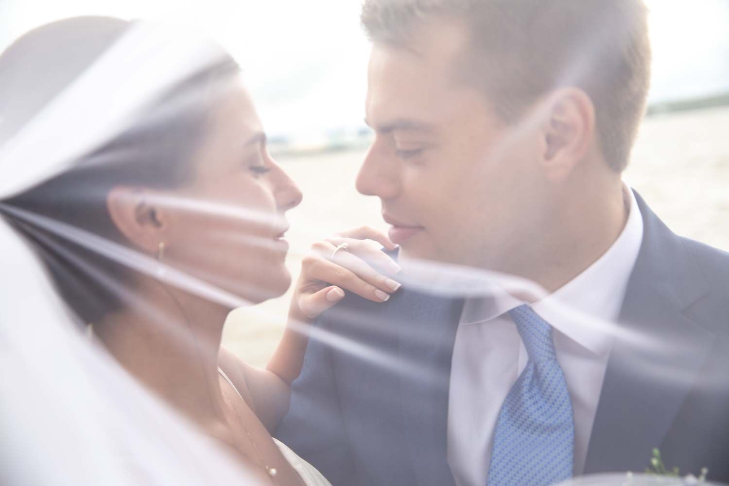 Intimate close-up of bride and groom almost kissing, softly lit through flowing white wedding veil.