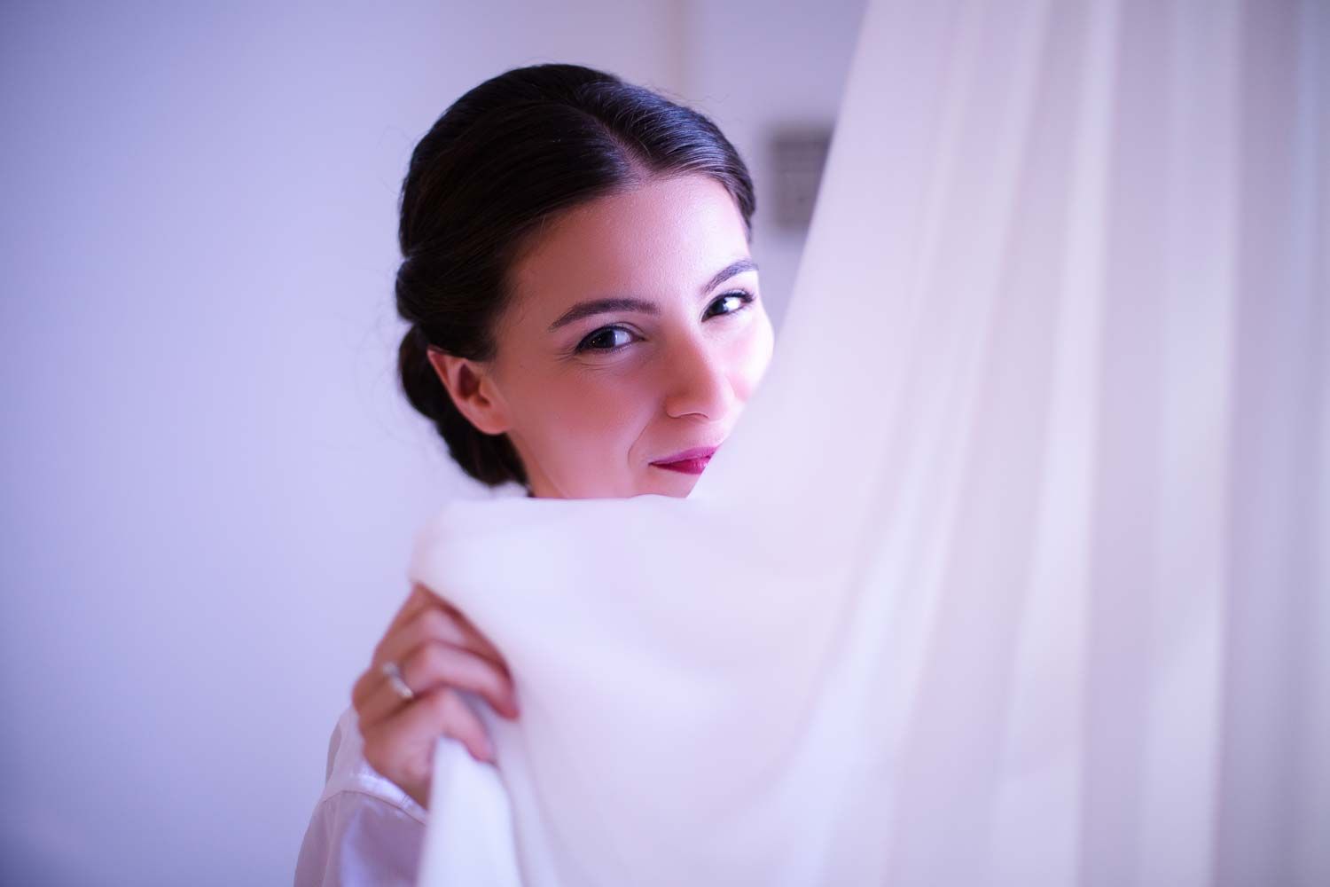 Beautiful bride with dark hair in an updo gazing over her shoulder through a flowing white curtain.