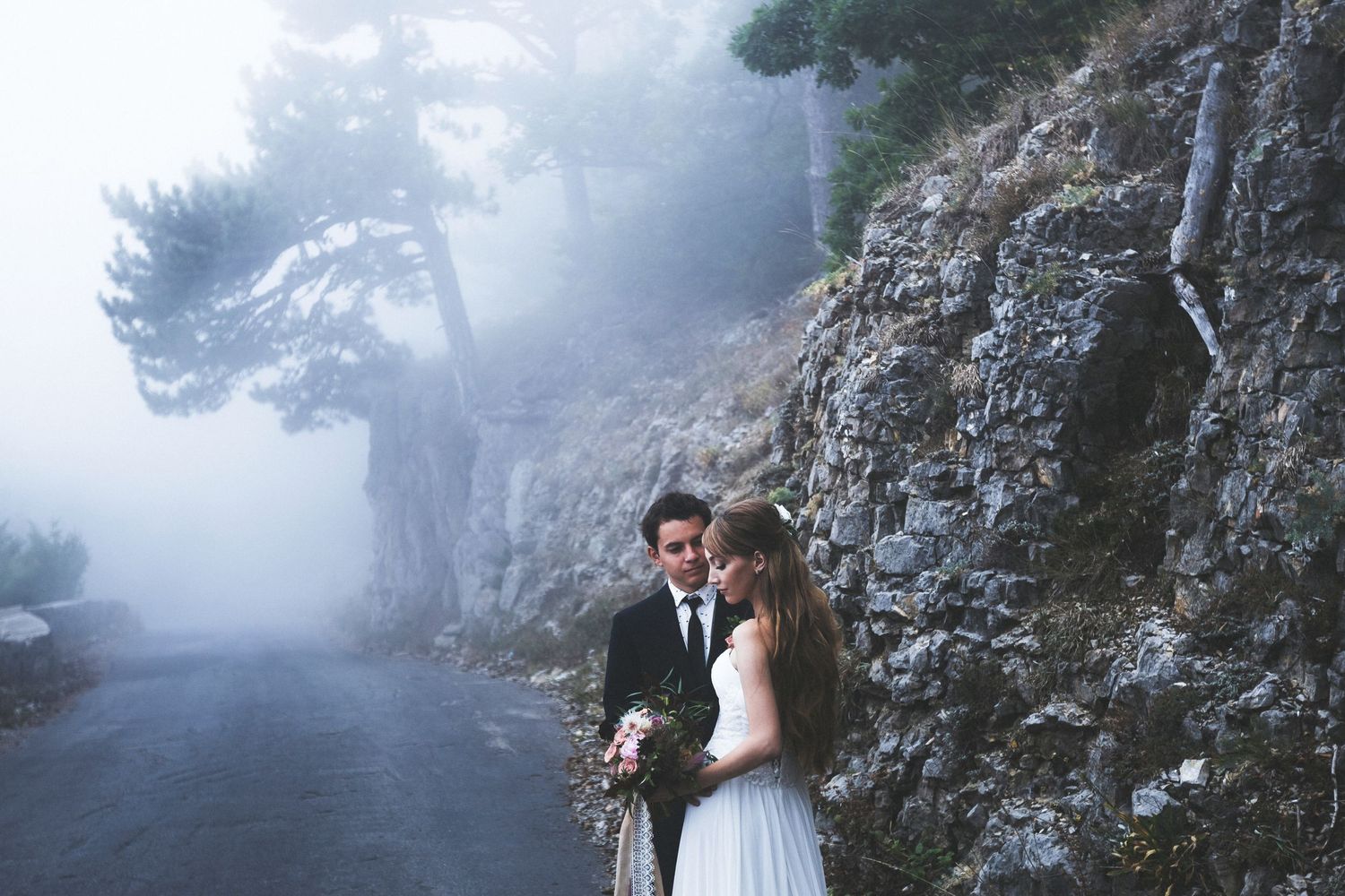 Bride and groom share a romantic moment on a misty mountain road surrounded by fog and rocky cliffs.