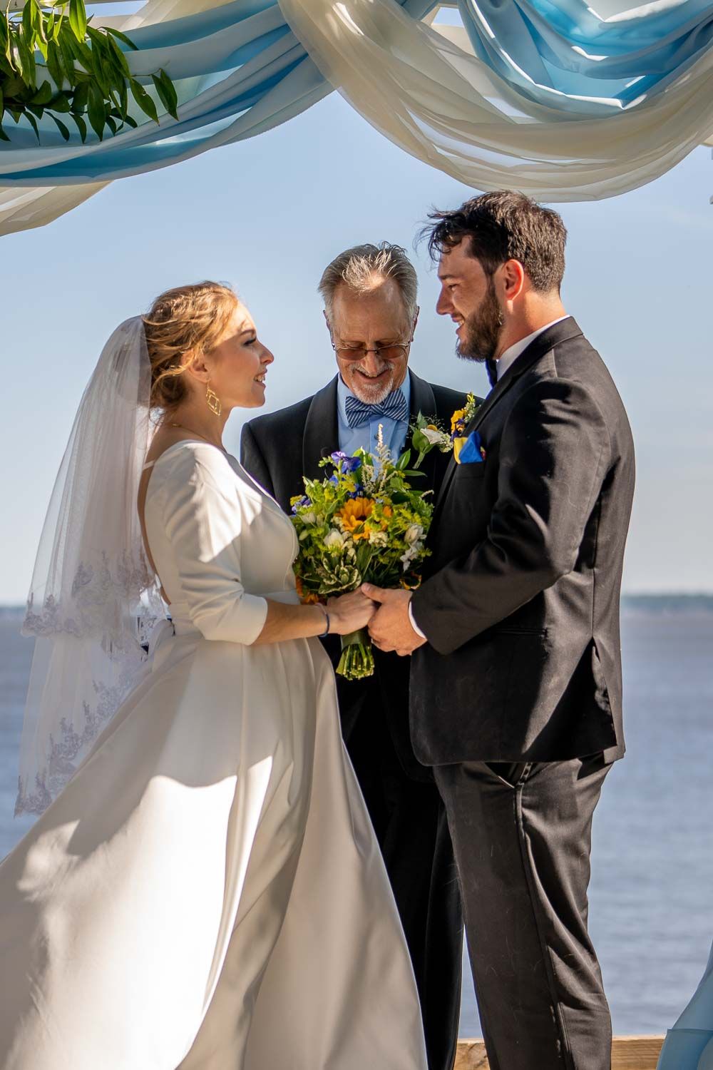 Bride and groom hold hands smiling as officiant presides over sunny waterfront wedding ceremony.
