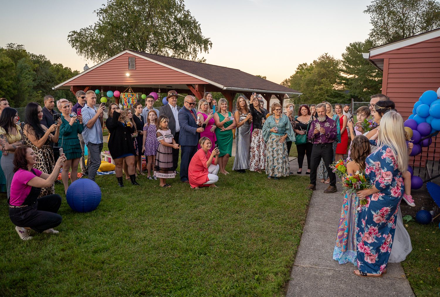 Wedding guests gather on lawn at dusk near decorated outdoor venue with colorful balloons and festive decorations.