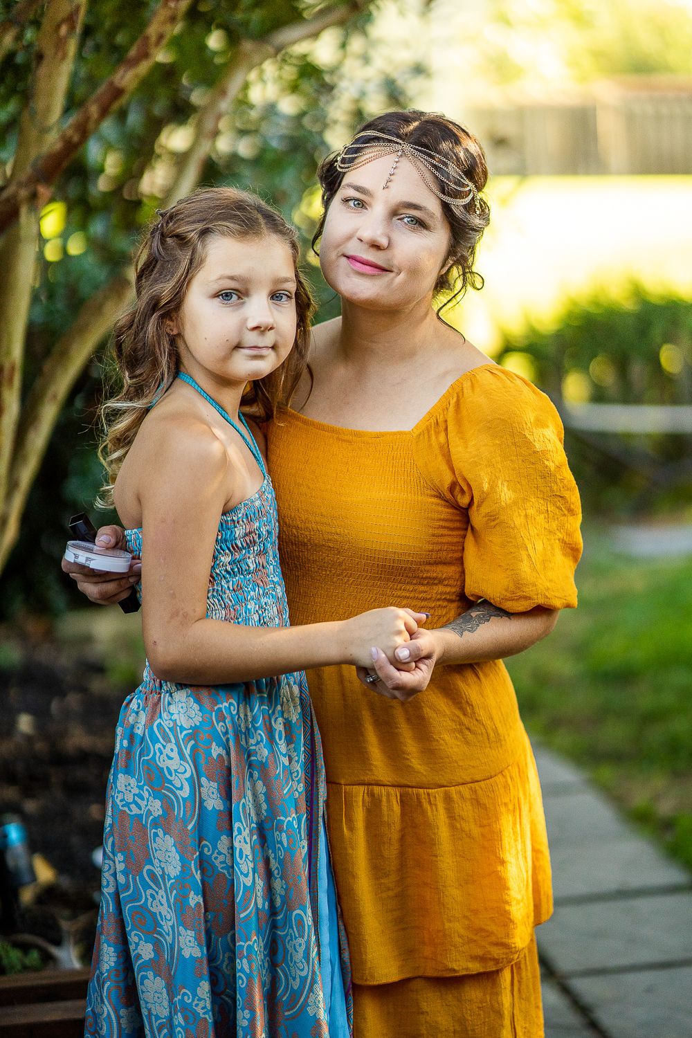 Woman in yellow dress with gold headchain hugging young girl in blue floral dress outdoors.