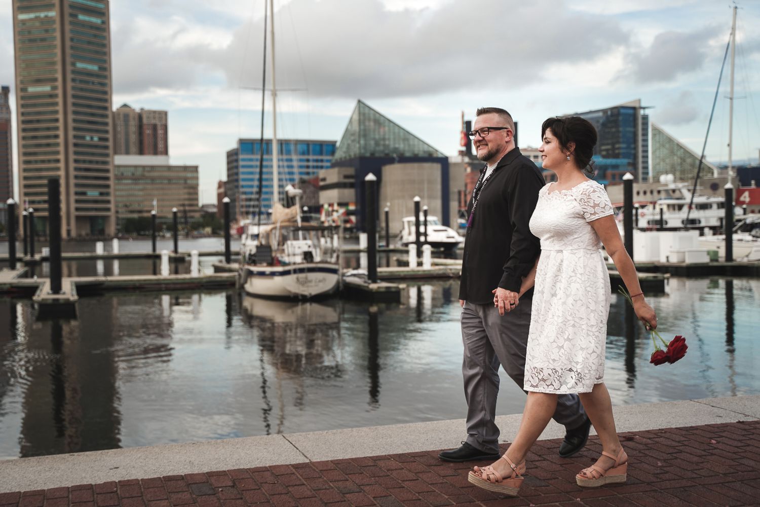 Newlyweds strolling along the Inner Harbor waterfront, bride in white lace dress holding red roses, groom beside her.