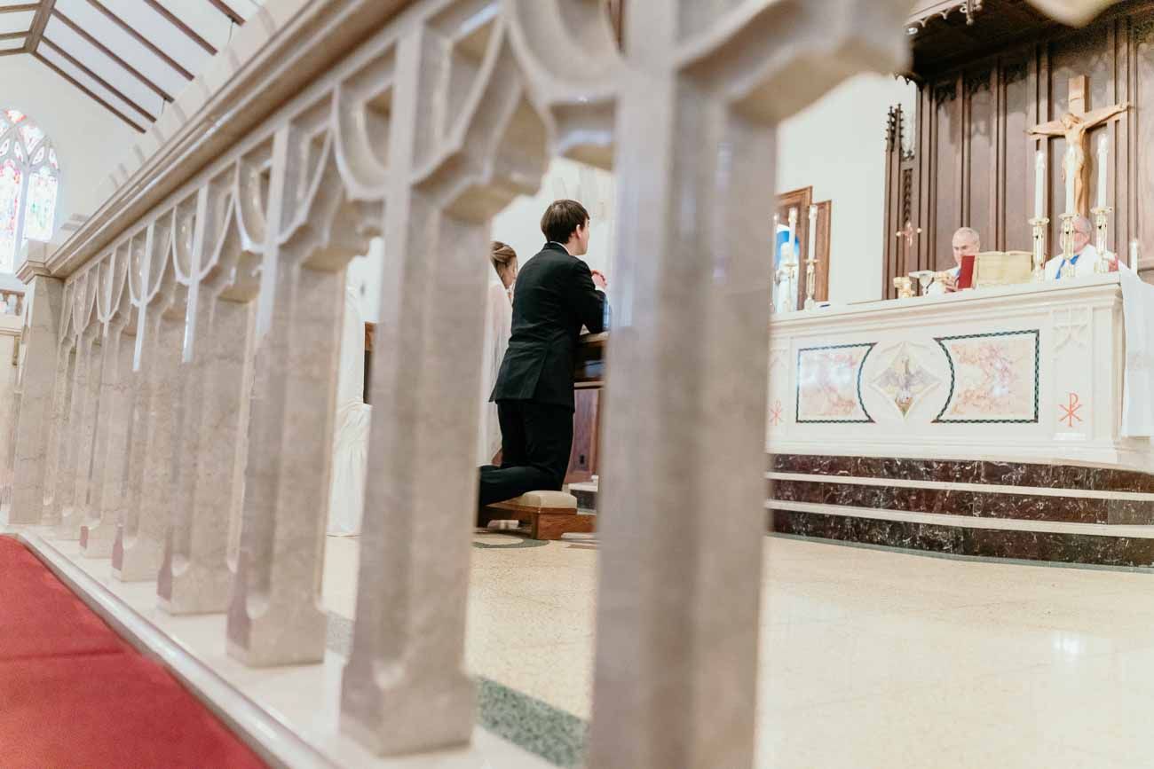Groom kneels at ornate marble altar rail during a traditional Catholic wedding Mass ceremony.
