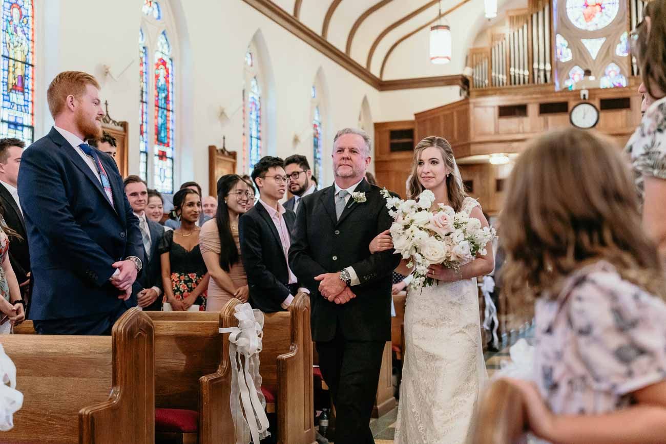 Father escorts bride in white gown down decorated church aisle toward waiting groom.
