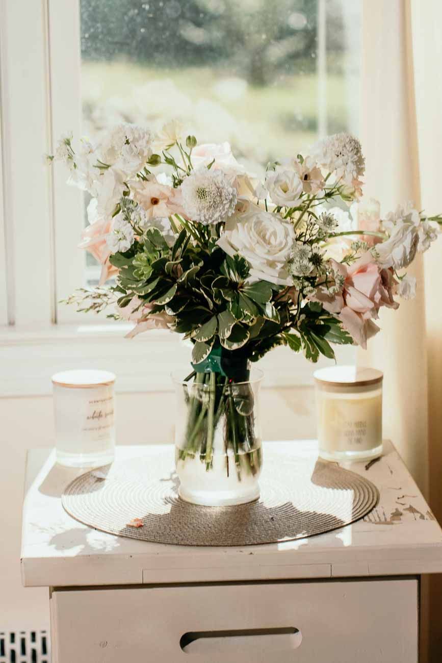 Elegant bridal bouquet of white and blush roses in glass vase on white table by sunny window.