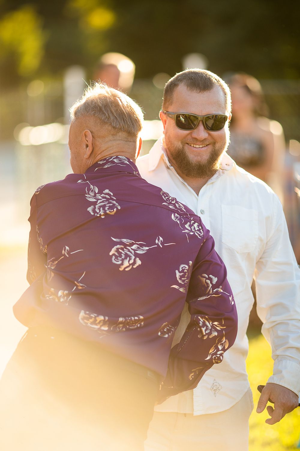 Smiling bearded man in sunglasses embracing an older man in a floral purple shirt at an outdoor event.