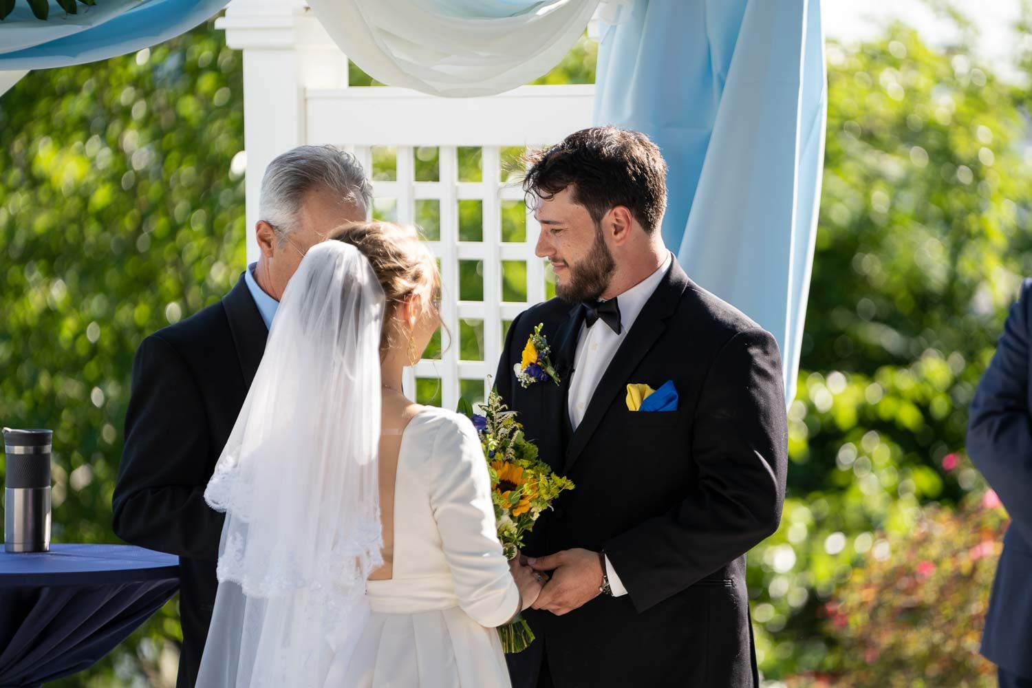 Groom smiles warmly at bride during outdoor wedding ceremony beneath white lattice arch with blue draping.