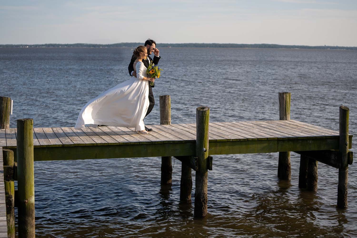 Bride and groom kiss on wooden dock overlooking calm blue water on their wedding day.