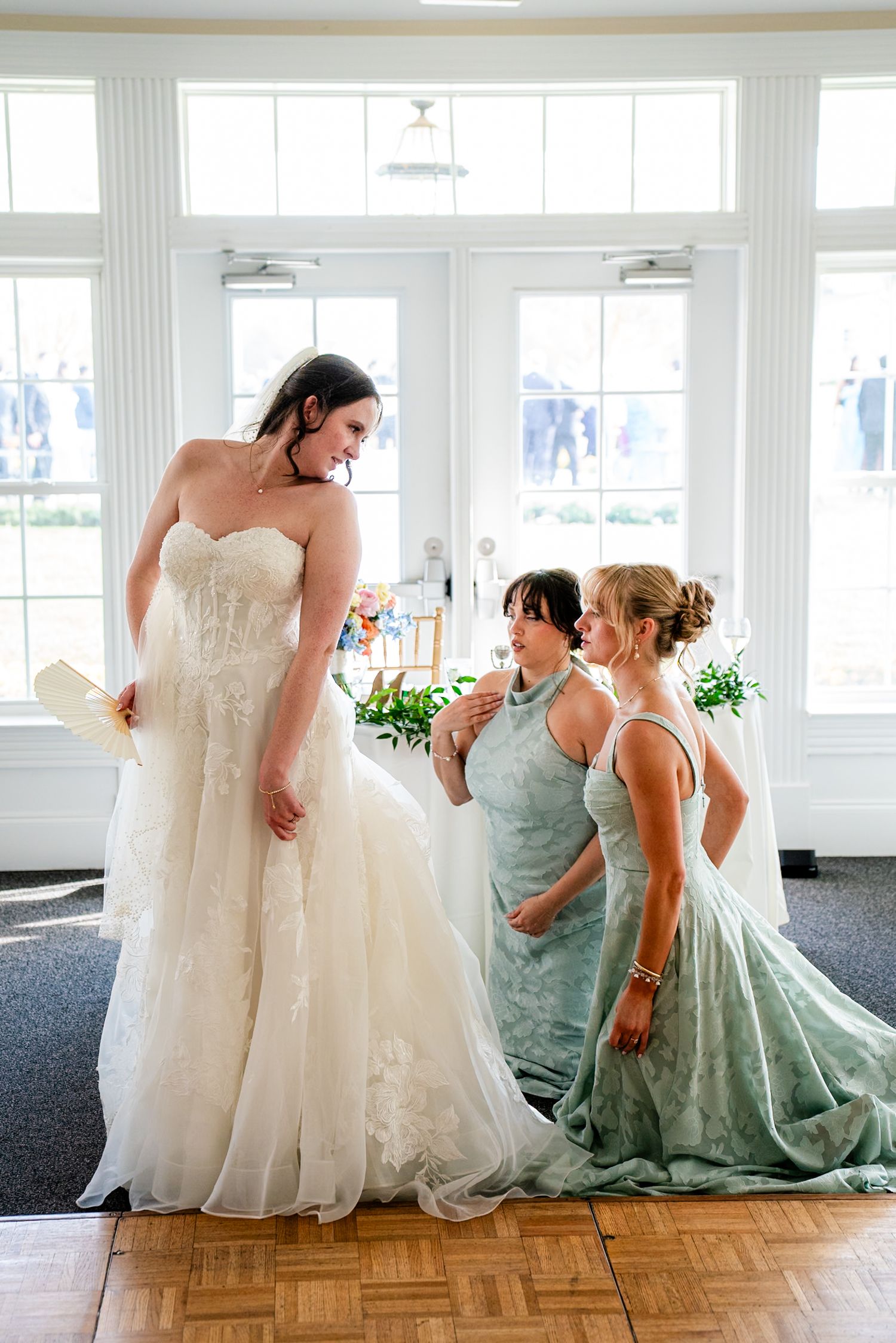 Bride in white lace gown with two women in sage green dresses helping her prepare in a bright sunlit room.