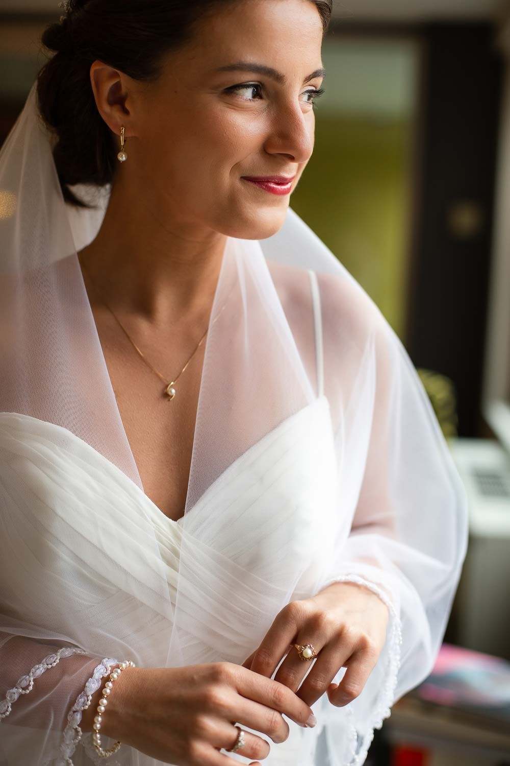 Radiant bride in white gown with pearl jewelry and veil smiling softly in natural window light.