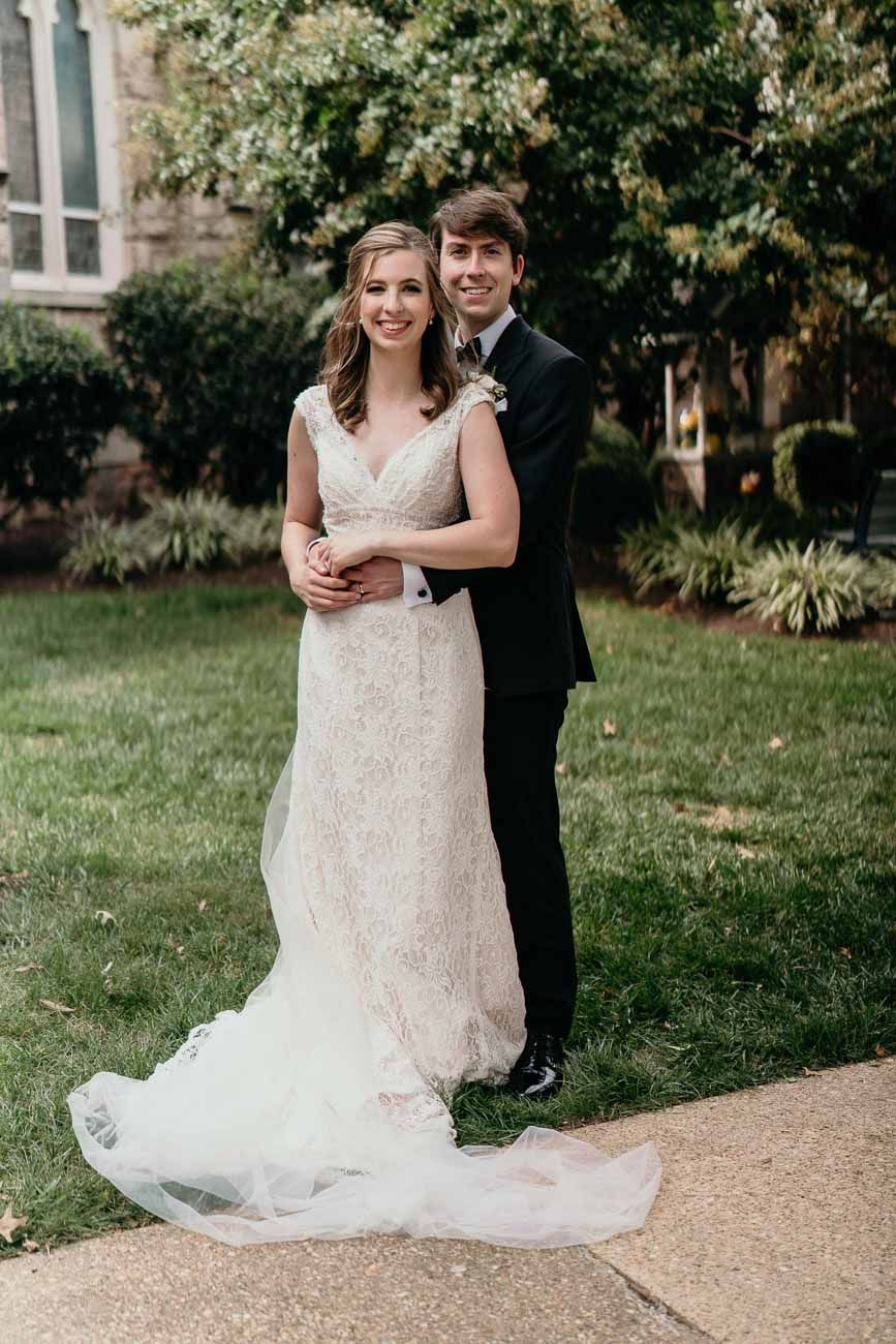 Groom embraces bride from behind as she holds a bouquet in a lace wedding gown on a church lawn.
