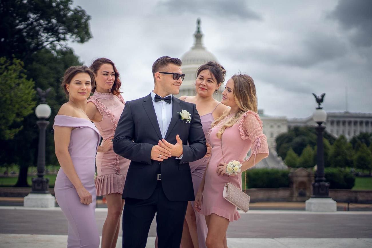 Groom in black tuxedo surrounded by bridesmaids in pink dresses near US Capitol.