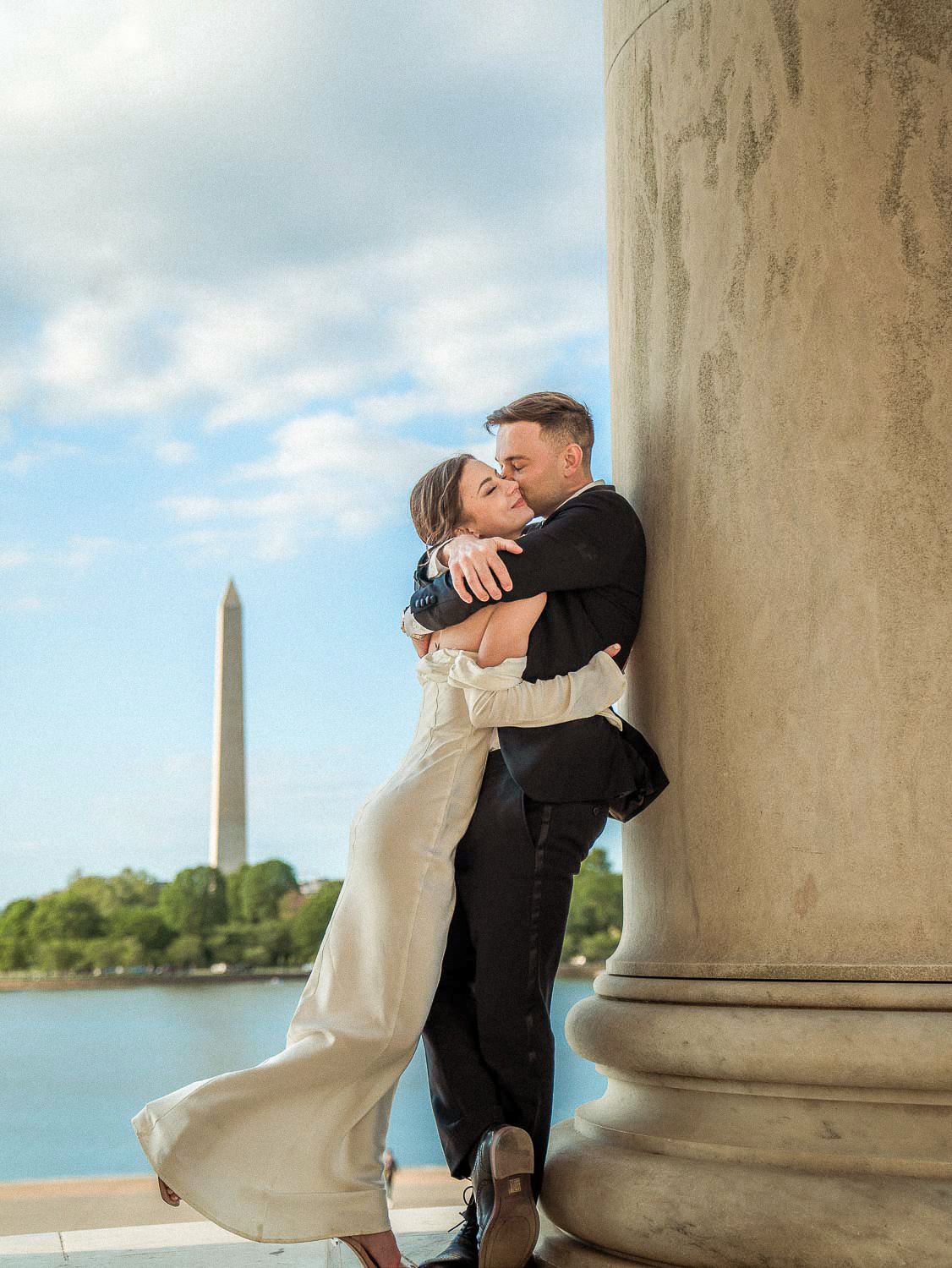 Bride and groom embrace passionately by a marble column with the Washington Monument in the background.